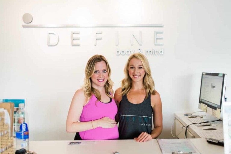 Two women stand at the reception desk of Define body & mind. The woman on the left, wearing a pink top, is pregnant and smiles warmly at the camera while resting her hand on her belly. The woman on the right, in a grey tank top, also beams at the camera.