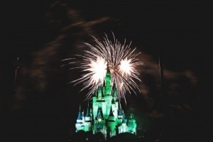 A brightly lit Disney castle, illuminated in green, stands against a dark night sky with fireworks bursting above it. The fireworks display creates a festive ambiance around the magical castle.