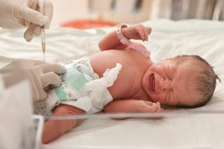 A newborn baby is lying on a hospital bed, crying as a medical professional wearing gloves prepares to administer a vitamin K shot at birth. The baby, donning a diaper and hospital identification wristband, is in a typical medical facility setting.