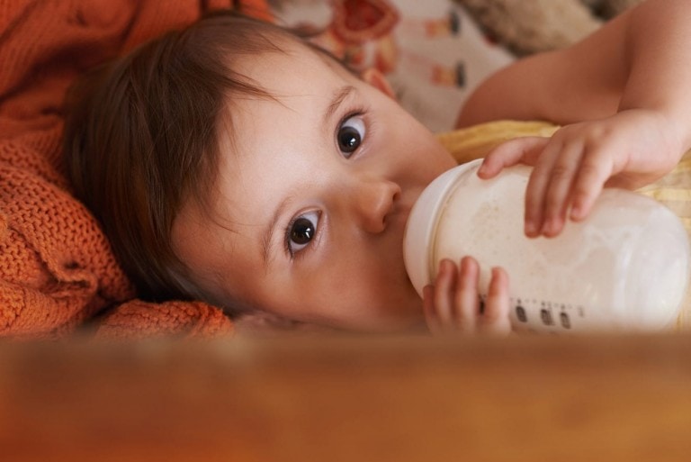 An infant lies on a patterned blanket, drinking from a baby bottle filled with formula. The baby, with brown hair and wide eyes, looks directly at the camera. An orange knitted blanket is partially visible in the background.
