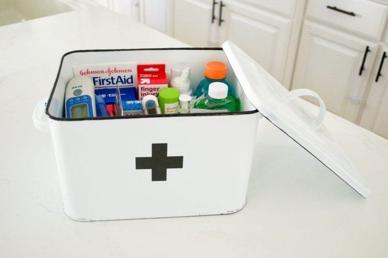 A white first aid box with a black cross on the front is shown with the lid open. Inside, various medical supplies are visible, including a thermometer, bandages, antiseptic wipes, and medications. This toddler's first aid kit is placed on a white countertop with cabinets in the background.