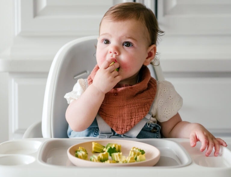 A baby sits in a high chair, wearing a bib and holding a piece of food. In front of the baby is a plate with small pieces of avocado, promoting healthy eating habits. The baby appears to be eating, with one hand holding food to its mouth. The background features white cabinetry.