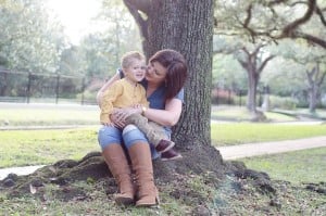 A woman sits under a tree holding a young boy on her lap. They are both smiling. The woman is wearing blue jeans and brown boots, and the boy is in a yellow shirt with beige pants. It's as if she's crafting a living letter to my son in this serene park with green grass and trees in the background.
