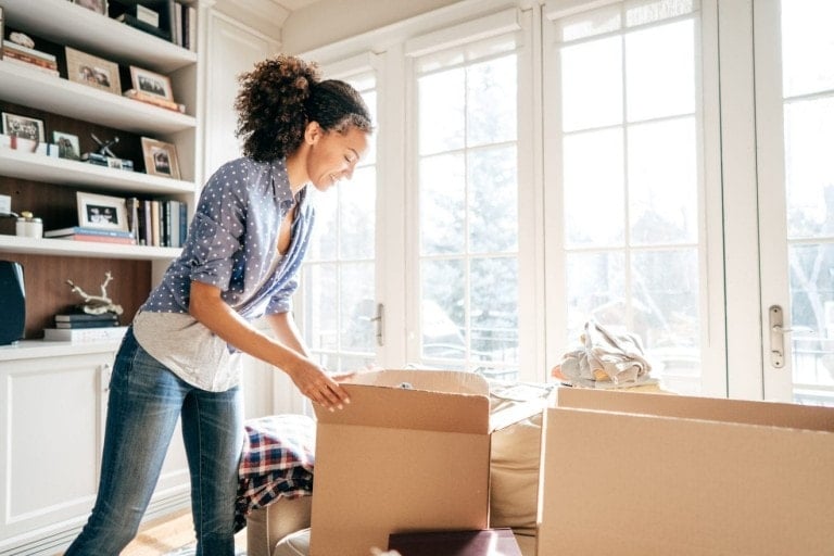 A woman is packing items into cardboard boxes in a well-lit living room with large windows and a glass door, getting her home organized. She is wearing a blue polka dot shirt and jeans. Shelves with pictures and books are visible in the background.