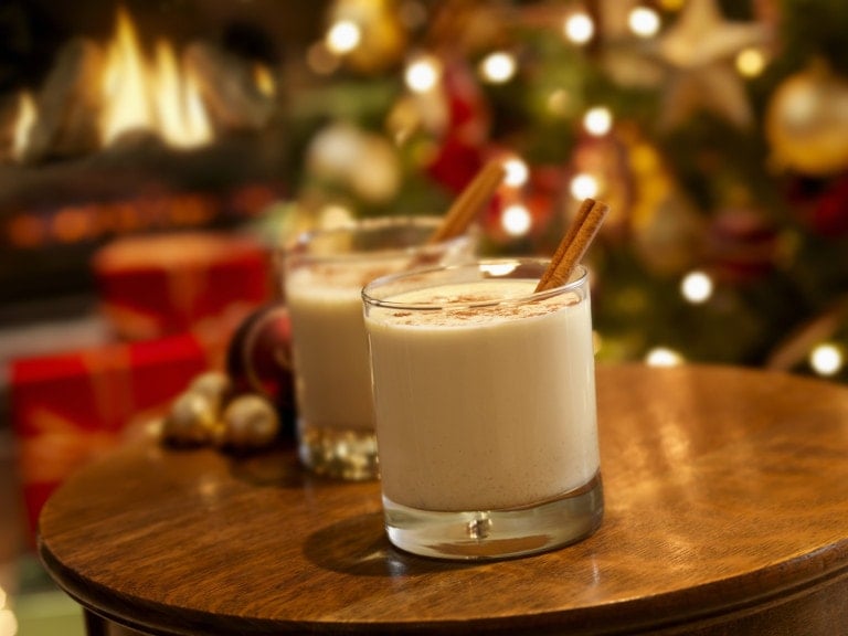 Two glasses of creamy holiday eggnog mocktail topped with cinnamon sticks sit on a round wooden table. In the background, there is a decorated Christmas tree with lights, ornaments, and a fireplace, contributing to a festive atmosphere.