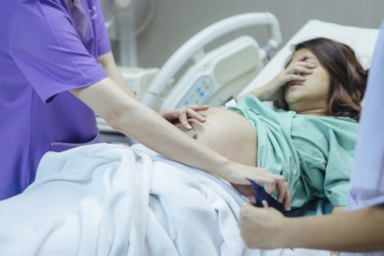 A pregnant woman lies on a hospital bed, covering her face with her hand. A medical professional in a purple uniform examines her abdomen with a device, while another person in white assists. The setting appears to be a labor and delivery room where the labor has stalled out.