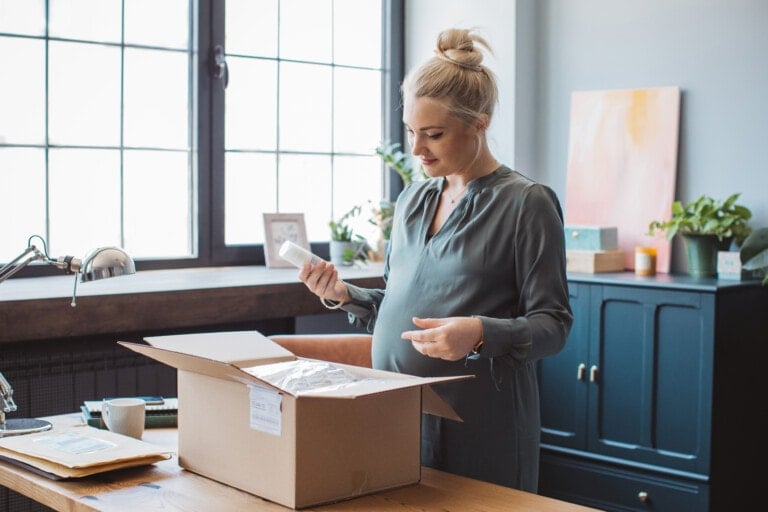 Portrait of pregnant woman received package, unpacking cardboard box. She is in living room at her apartment. Happy with online purchase