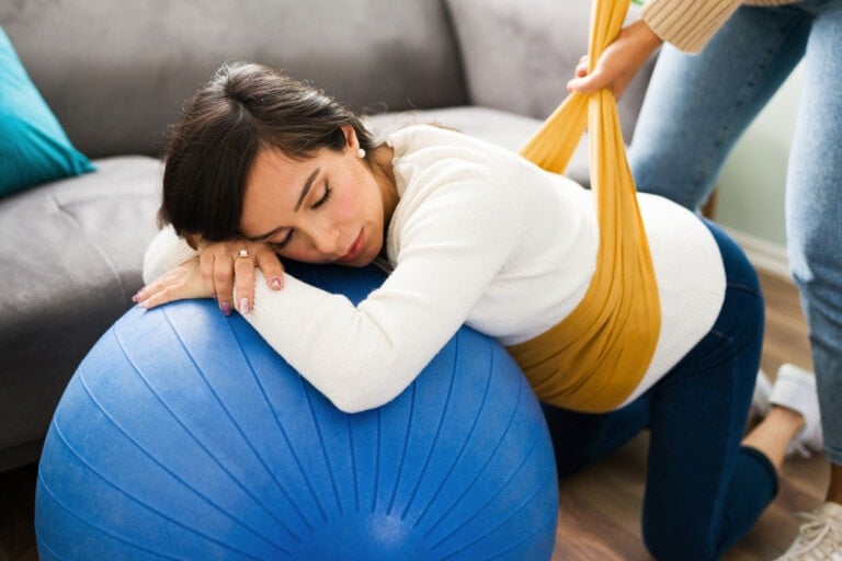 A woman leans forward with her eyes closed on a blue birthing ball while another person holds a yellow rebozo wrap around her upper torso. She is in a home setting with a couch visible in the background.