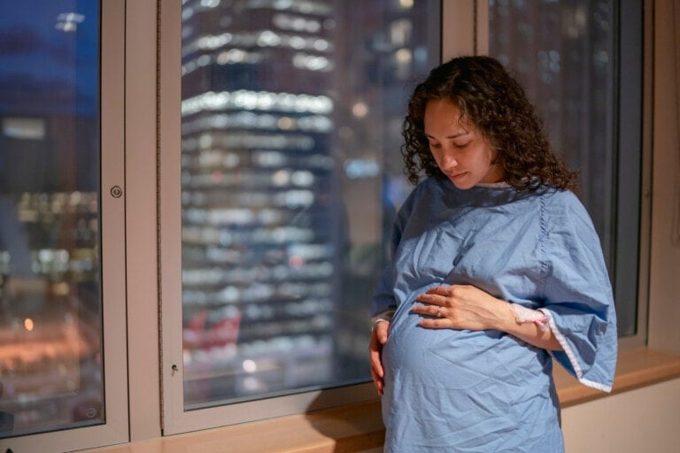 A pregnant woman wearing a hospital gown stands by a window, looking down and gently holding her belly. The window shows the blurred lights of a cityscape at night, as she contemplates not just the future but also the cost of having a baby.