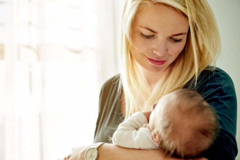 A woman with long blonde hair is holding an infant in her arms near a sunlit window. She looks down at the baby with a gentle expression. Wearing a watch and a dark top, she epitomizes self-care for moms, ensuring both of them are enveloped in love as the baby is wrapped in a light-colored blanket.