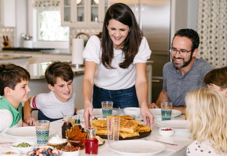 A woman, known as The Baker Mama, is serving a stack of pancakes to a family seated around a table. The table is set with plates, glasses filled with water, and various breakfast items including syrups, berries, and whipped cream. Four children and one man are seated at the table.