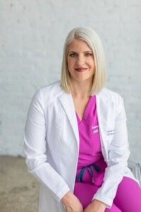 A woman with shoulder-length blonde hair is wearing a white lab coat over a pink medical outfit. She is seated against a light gray brick wall, looking at the camera with a slight smile.
