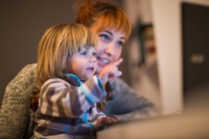 A small child with blonde hair and a striped sweater points at a computer screen while sitting on the lap of an adult with orange hair, who is also looking at the screen. Both appear to be inside a dimly lit room, possibly watching educational shows for toddlers together.
