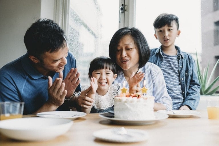 A family of four sits around a table celebrating a young girl's birthday. The girl, positioned between her parents, smiles while her mother helps her blow out candles on a white frosted cake topped with strawberries. A boy stands and looks on from behind, highlighting how easy it is to simplify kids' birthday parties.