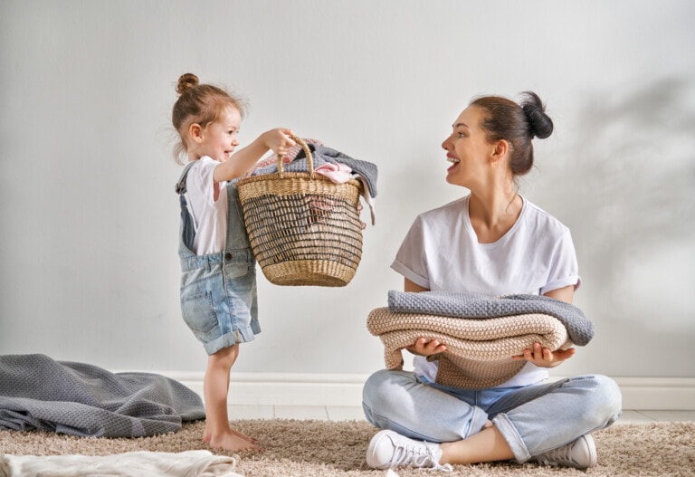A smiling woman is sitting on the floor, holding a pile of folded laundry. A young child, also smiling, stands nearby, holding a woven basket filled with clothes. Both are engaged in a light-hearted moment, likely related to household chores and sharing cleaning tips along the way.