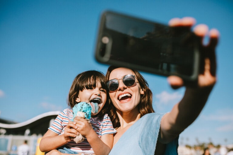A woman wearing sunglasses takes a selfie while holding a child. The child, eating a scoop of blue ice cream, beams with joy. They are both smiling in the picture-perfect moment against a backdrop of blue sky and architectural structures, embracing the essence of sharenting.