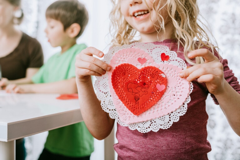 A young child holding a handmade heart-shaped craft made of red and pink paper, adorned with smaller hearts. The child is smiling, and there are two other children in the background focused on their own crafts at a table—perfect class valentine ideas for Valentine's Day.