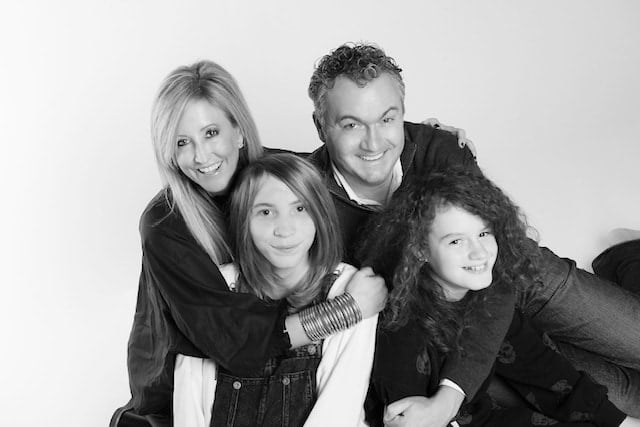 Black and white photo of a family of four on their adoption journey. An adult woman and man are in the back, smiling, with the woman leaning forward. Two children, one with straight hair and the other with curly hair, are in front, also smiling. They are positioned closely together.