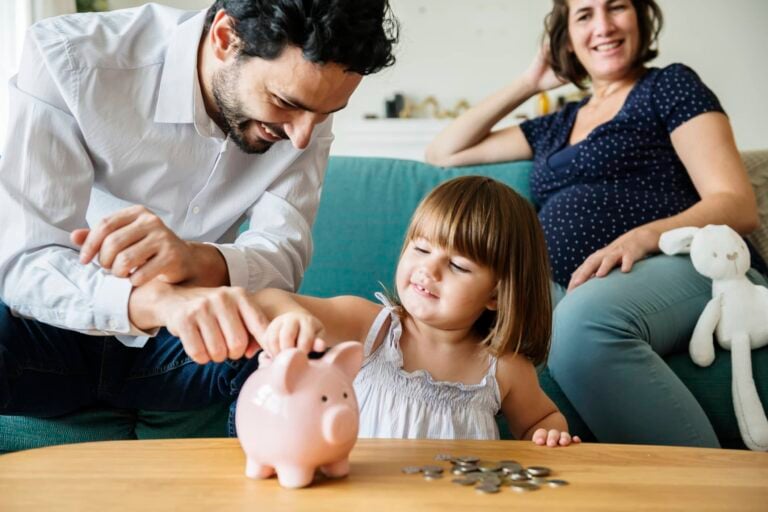 A young girl is placing coins into a pink piggy bank, building good money habits, while her father smiles and helps her. They are seated on a light blue couch, and a woman is sitting comfortably beside them with a white stuffed animal in the background.