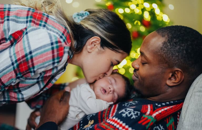 Amidst the holidays with a newborn, a woman kisses her baby held by her parnter sitting beside her, near a twinkling Christmas tree. She wears a plaid shirt, he sports a festive sweater, and their tiny baby is wrapped snugly in a light-colored blanket.