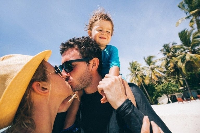 A couple kisses passionately while standing on a sunny beach, trying to get the passion back. The man is holding a young child on his shoulders. The woman wears a hat, and the man sports sunglasses. Palm trees, blue sky, and lounge chairs are visible in the background.