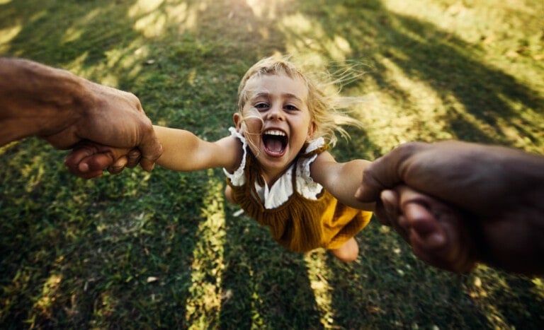 A young child, smiling widely, is being swung around by two adults holding her hands. She is wearing a yellow dress and appears to be enjoying the playful activity in an outdoor grassy area—one of many wonderful things to do while your child is little.