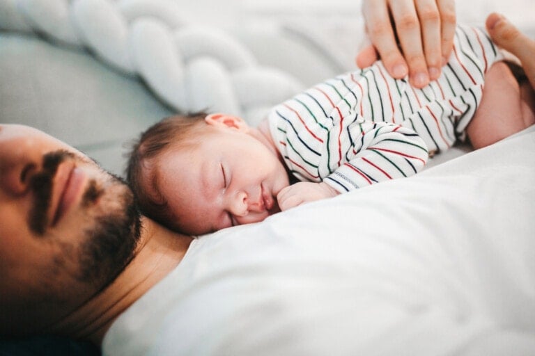 A baby wearing a striped onesie sleeps on the chest of an adult who is lying down. The adult's face is partially visible, and their hand is gently placed on the baby's back. The partner's presence makes the scene feel special, exuding a calm and intimate ambiance.