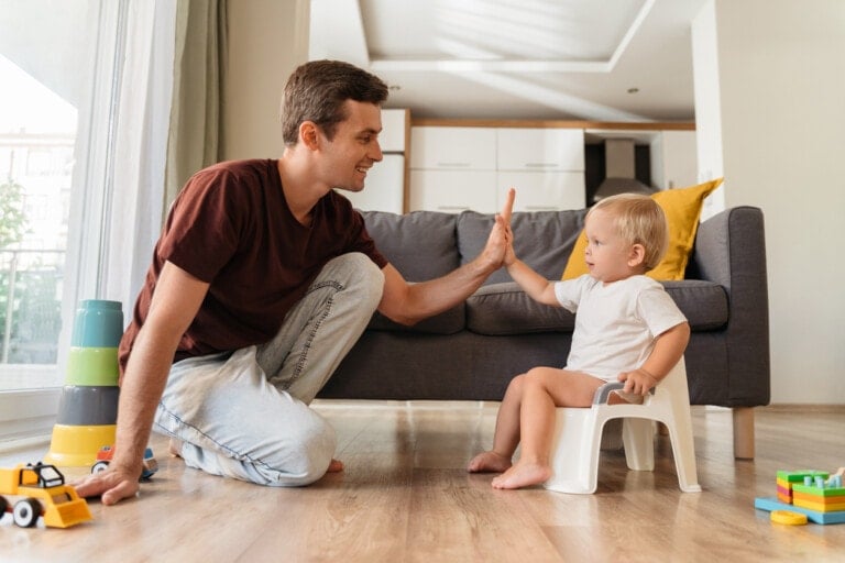 A man kneels on the floor, giving a high-five to a young child sitting on a small potty in the living room. Colorful toys are scattered about, and a gray couch with a yellow pillow is in the background. The room is well-lit with natural light. This scene perfectly illustrates successful potty training tips in action.