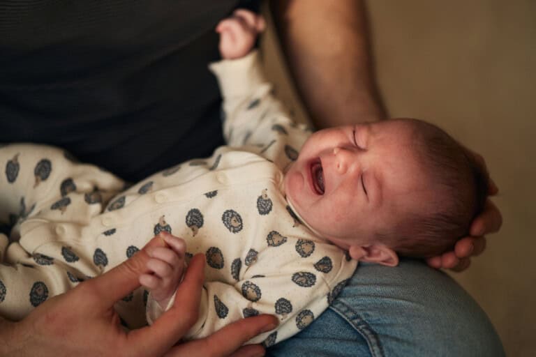 A baby wearing a white onesie with a hedgehog pattern is crying while lying on an adults lap. The adults hand is gently cradling the babys head, perhaps worried about dehydration in infants. The background is out of focus.
