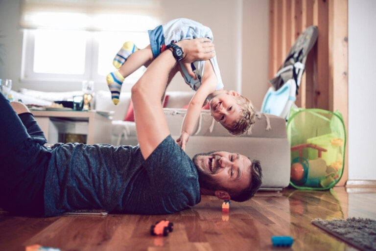 A man lying on a wooden floor enjoys playtime with his smiling child, holding them upside down above him. The room is strewn with colorful toys, and a beige sofa with cushions sits in the background. A green mesh basket filled with toys is visible to the right.