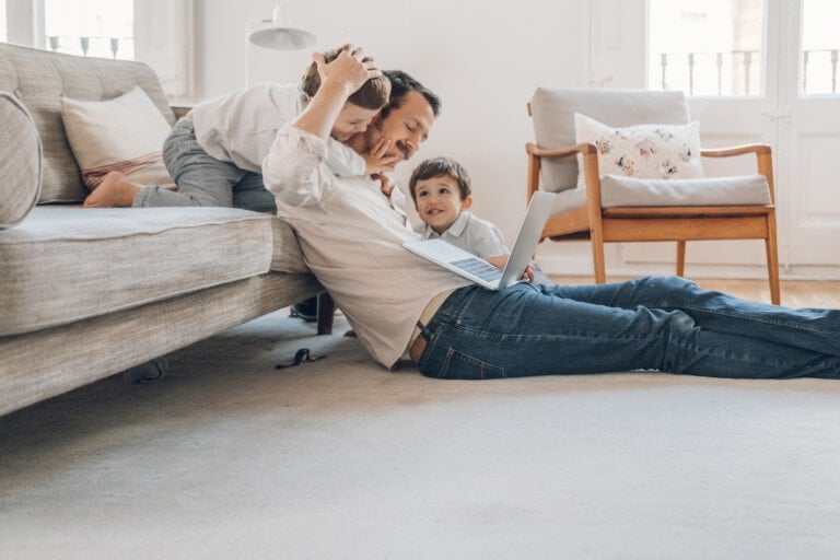 A man lies on the floor using a laptop, while a child hugs him from behind on the sofa, and another child sits beside him smiling. The room's light-colored interior features a chair and a cushion in the background, creating a cozy atmosphere that encourages everyone to behave better.