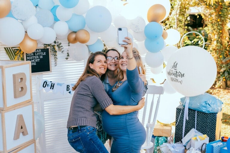 Three women pose for a selfie at a baby shower, standing in front of a balloon arch adorned with white, blue, and gold balloons. One woman holds the phone while the other two embrace her. Surrounded by gifts and decorations, including a large white balloon that reads "Let the adventure begin," they look ready to capture memories for the baby shower invitation.