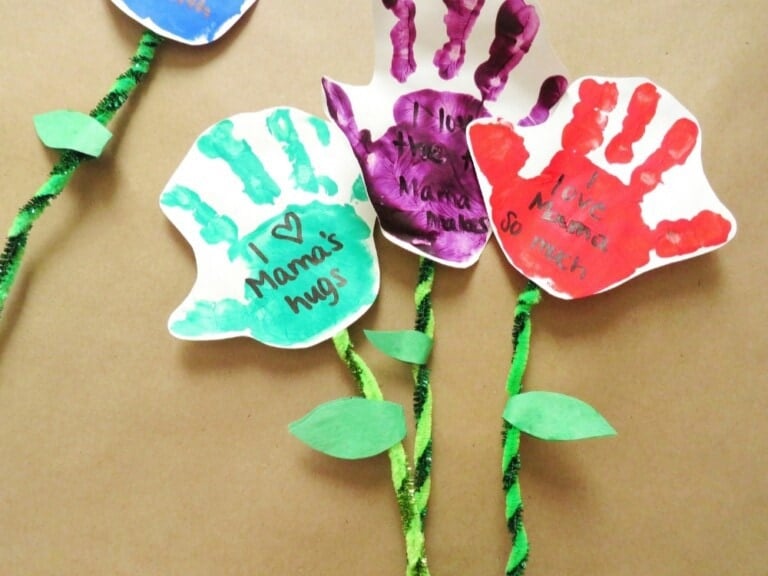 Three colorful paper flowers made from handprints on white paper make a delightful Mother's Day craft. The handprints are painted green, purple, and red, each carrying a heartfelt note expressing love for "Mama." The stems are green pipe cleaners adorned with green paper leaves.