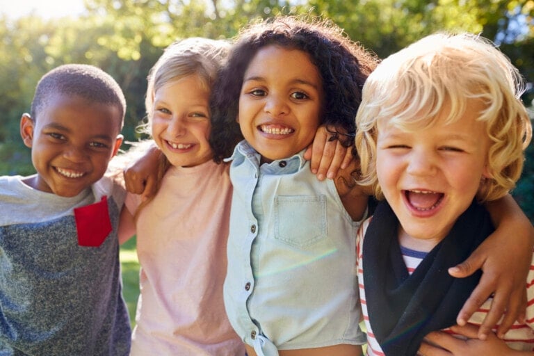 Four children stand outdoors with their arms around each other, smiling broadly at the camera. They are in a park or garden setting with green trees in the background, embodying perfect summer playdate ideas. The children are wearing casual clothes and appear to be enjoying a sunny day.