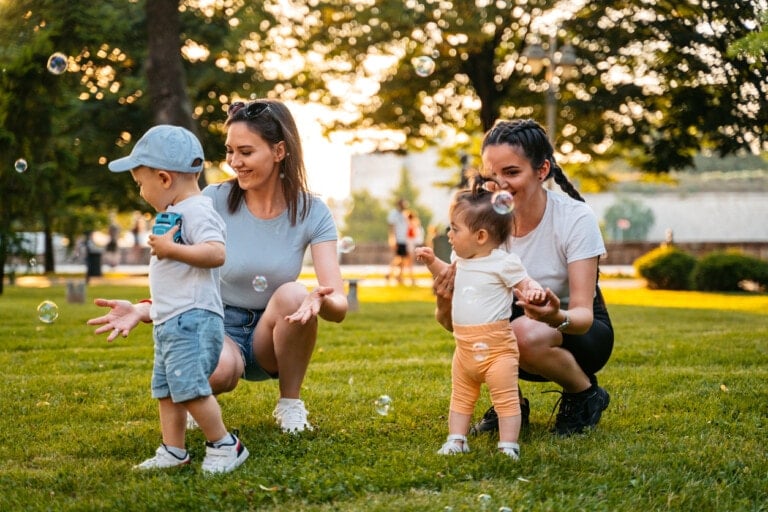 Two women and two toddlers are enjoying a playdate in a grassy park, delighting in the magic of bubbles. The toddlers stand while the women kneel, fully engaged with the children's joy. Sunlight filters through trees in the background, casting a warm glow on the scene as bubbles float around them.