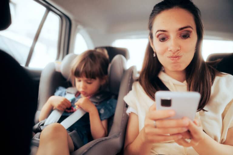 A woman sits in the back of a car looking at her smartphone, making a funny face—clearly one of those modern mom probs. Next to her, a young girl, her daughter, is seated in a child car seat, focusing intently on a toy as both enjoy their journey.