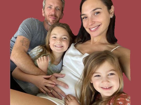 A man, woman, and two young girls are smiling at the camera. The woman, reminiscent of Gal Gadot pregnant, is wearing a white dress and has her hand on her belly. One girl is also touching the belly, while the man is embracing the woman from behind. They are against a plain red background.
