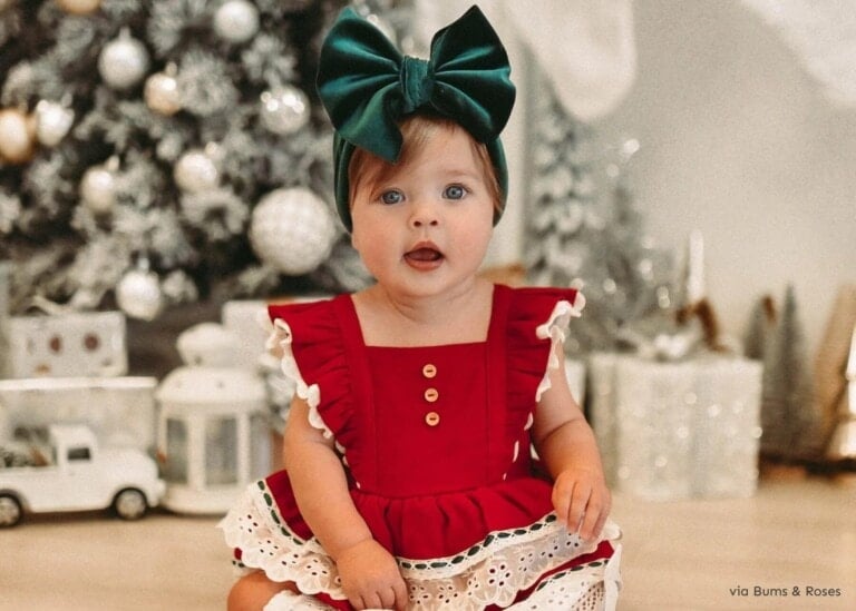 A baby in a cute Christmas outfit—specifically, a red dress with white lace and a large green bow headband—sits in front of a decorated tree. The toddler looks directly at the camera, with holiday decorations and gifts beautifully arranged in the background.