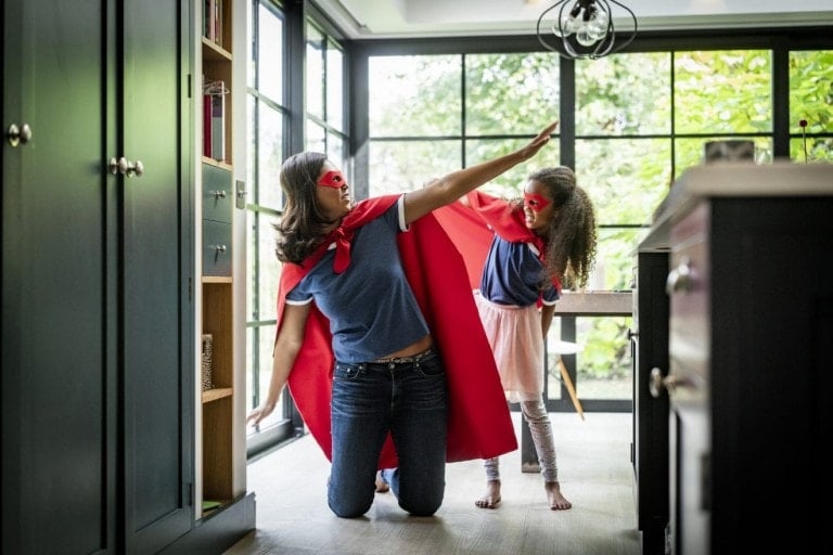 A playful parent and a child, both wearing red capes and red masks, are pretending to be superheroes in a modern kitchen with large windows. The parent kneels on one knee with an arm raised forward, while the child stands behind with an arm outstretched.