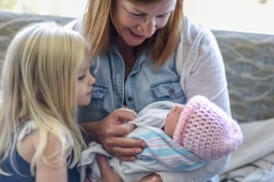 A woman, wearing a light blue shirt, holds a newborn baby wrapped in a striped blanket and wearing a pink knitted hat. An older child with long blonde hair looks at the baby while visiting a new baby. They are sitting on a couch.