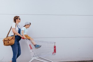 A woman pushing a shopping cart with a young boy sitting inside is saving time grocery shopping. The woman is carrying a tan handbag and wearing sunglasses, a white shirt, and overalls. The boy is sporting a gray hat, blue shirt, brown shorts, and blue shoes. They are walking past a white wall.