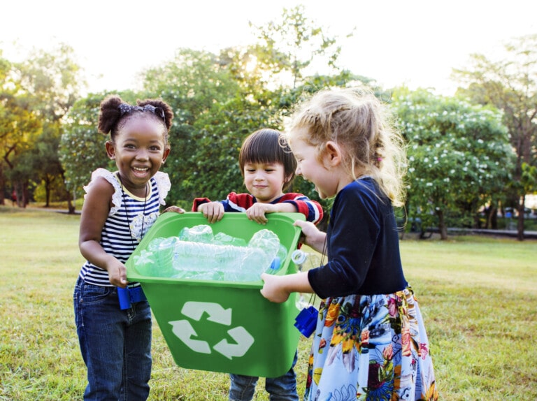 Three children are outdoors, smiling behind a green recycling bin filled with plastic bottles. They are standing on grass with trees and foliage in the background, showcasing one of the easiest volunteer ideas for kids—recycling.