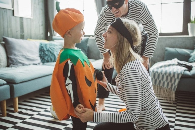 A child in a pumpkin costume smiles at an adult holding their hands, while another adult wearing a mask and striped shirt looks on, also smiling. They are indoors in a living room with a striped rug and a blue couch, enjoying their family Halloween costumes together.