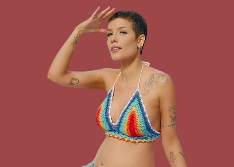 Person with short hair wearing a colorful, crocheted halter top, posing against a plain red background. They have tattoos on their arm and torso and are gazing into the distance with one hand raised to their forehead, reminiscent of Halsey gives birth to new beginnings.