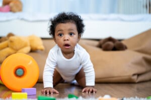 A baby boy wearing a white onesie is crawling on a wooden floor. The baby, who might someday be exploring boy names that start with X, has curly hair and is surrounded by colorful toys, including a large orange ring and some foam blocks. There are plush animals and a bed in the background.