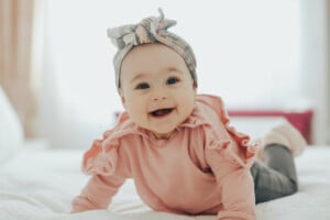 A baby with a happy expression is lying on their stomach on a white bed. The baby, who could be one of the charming F girl names, is wearing a pink ruffled top, grey pants, and a grey headband with a bow. The background is softly blurred, focusing on the baby's smiling face.