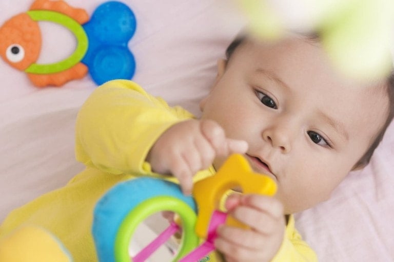A baby wearing a yellow outfit lies on a soft surface, holding and examining a colorful sensory toy with rings and shapes. A fish-shaped toy with blue and orange sections is visible in the background. The baby appears focused on the sensory toy for toddlers.
