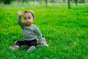 A toddler named Ella sits on the grass in a park, wearing a patterned long-sleeve shirt, black pants, and pink shoes. The child has a headband and is holding one hand up. Trees and greenery are in the background.