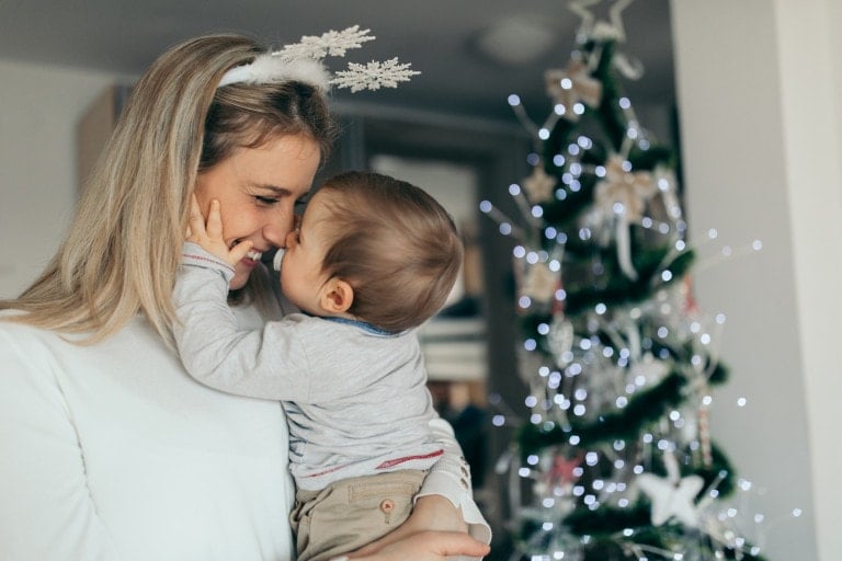 A woman wearing a white outfit and snowflake headband holds a baby close, touching noses. They are in front of a decorated Christmas tree with white lights and ornaments. The scene is indoors and festive, capturing the magic of the season—perfect for thinking about stocking stuffers for babies.