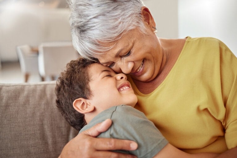 A woman with short gray hair and a yellow top is sitting and hugging a young boy with short brown hair. Both are smiling and closing their eyes, conveying a sense of warmth and affection. They are indoors on a beige couch with a blurred background, perfect for emphasizing the special bond in personalized gifts for grandparents.